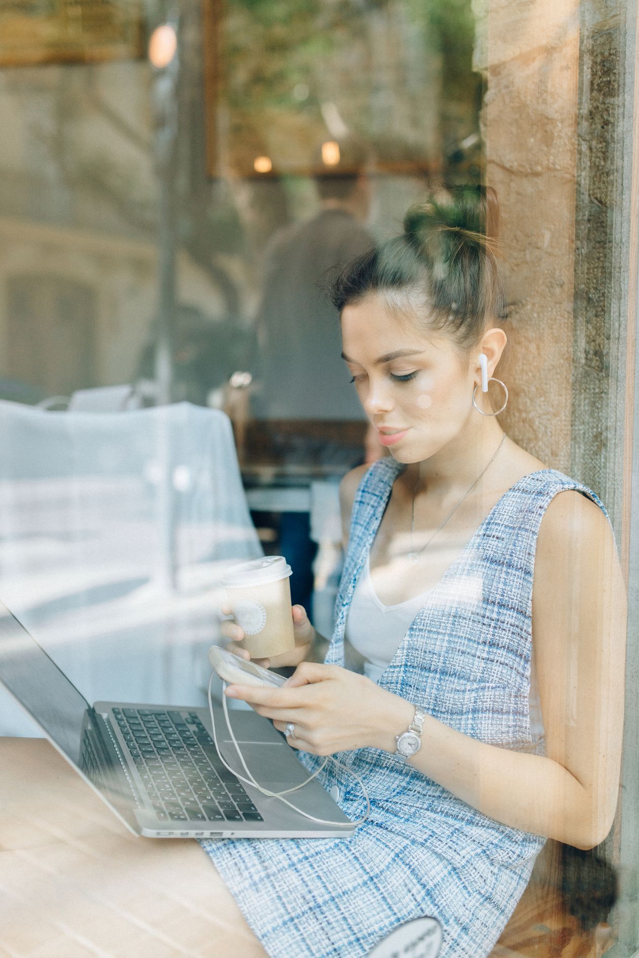 A woman sitting at a cafe window, using a laptop and phone.