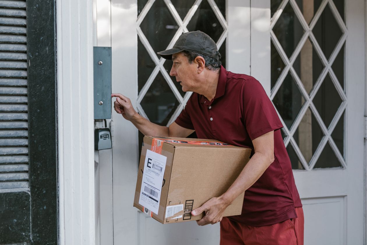 A delivery driver checking his phone beside a parcel at a front door with a video doorbell.