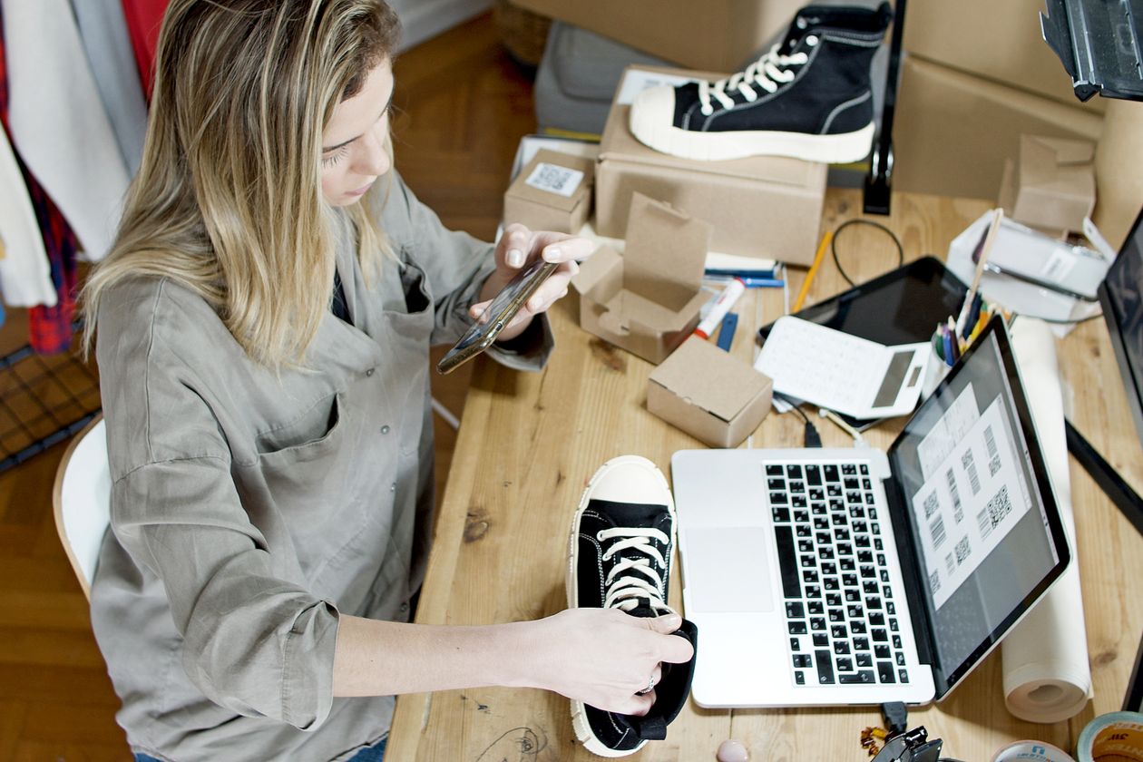 A small business owner photographing stock with a phone at a cluttered desk, laptop open beside her.